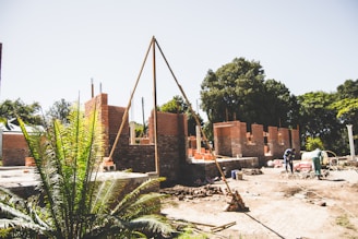An outdoor construction site with partially built brick walls and scattered building materials. The scene includes workers engaged in construction activities under a clear sky. A large brick structure is taking shape amidst trees and greenery, hinting at a rural or suburban setting.
