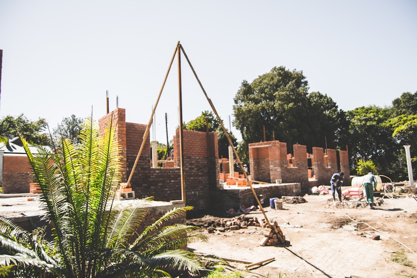 An outdoor construction site with partially built brick walls and scattered building materials. The scene includes workers engaged in construction activities under a clear sky. A large brick structure is taking shape amidst trees and greenery, hinting at a rural or suburban setting.