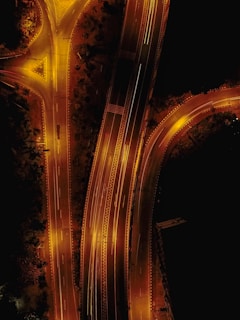 Aerial view of a well-lit highway interchange at night. The roads and lanes are illuminated by streetlights, casting a warm, golden glow. The highway splits into multiple directions, showing an intricate design of curves and intersections, surrounded by patches of darkness from the surrounding unlit areas.