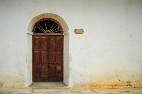 A rustic wooden door set into a weathered, whitewashed wall. The wall features peeling paint with hints of yellow and a small plaque beside the door bearing the text 'CASTILLO DE ARISMENDI'. The arched top of the door has decorative ironwork.