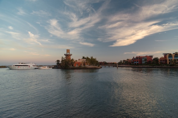 A serene waterfront scene featuring a large yacht and a small boat near an island with a lighthouse. The sky is partly cloudy with soft hues of blue and orange as day transitions to evening. Colorful buildings line the water's edge, surrounded by lush greenery.