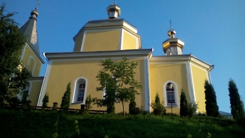 A yellow, orthodox-style church with golden domes rises against a clear blue sky. The structure is surrounded by small trees and shrubs, and the building features arched windows.