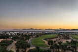 A panoramic view of a lush golf course with neatly manicured greens and palm trees. In the background, a distant mountain range silhouetted against an orange and grey sky at sunset. A cluster of buildings is visible at the edge of the course, adding an urban touch to the serene landscape.