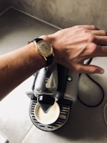 A close-up of a person's wrist wearing a classic wristwatch with a leather strap, positioned over a coffee machine that is dispensing coffee into a cup.