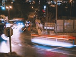 A nighttime urban scene with long exposure photography capturing light trails from moving vehicles at an intersection. The street is wet, reflecting various lights, and traffic signals are visible. A stone wall and some metal structures are on the right side, with trees and stairs in the background.