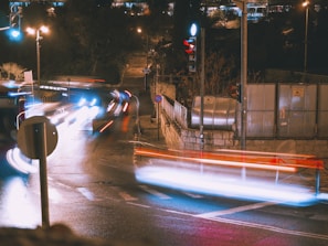 A nighttime urban scene with long exposure photography capturing light trails from moving vehicles at an intersection. The street is wet, reflecting various lights, and traffic signals are visible. A stone wall and some metal structures are on the right side, with trees and stairs in the background.