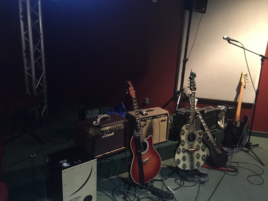 Two acoustic guitars are positioned in front of guitar amplifiers, surrounded by various other musical equipment including a microphone and a bass guitar. The room has dark walls, and the lighting creates a cozy and intimate atmosphere.