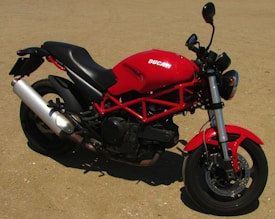 A red motorcycle with a sporty design is parked on a rough, sandy terrain. The bike features a black seat, prominent exhaust pipe, and rugged tires.