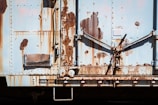 A weathered and rusty metal surface with visible signs of wear and age, featuring various shades of rust and peeling paint. The surface has mechanical elements, including levers and riveted panels.