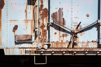 A weathered and rusty metal surface with visible signs of wear and age, featuring various shades of rust and peeling paint. The surface has mechanical elements, including levers and riveted panels.