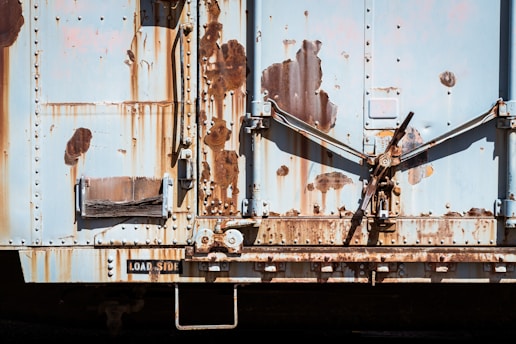 A weathered and rusty metal surface with visible signs of wear and age, featuring various shades of rust and peeling paint. The surface has mechanical elements, including levers and riveted panels.