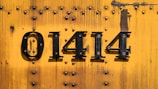 A vibrant yellow metal surface with multiple rivets, featuring the black painted numbers 01414. The background shows signs of wear and rust.