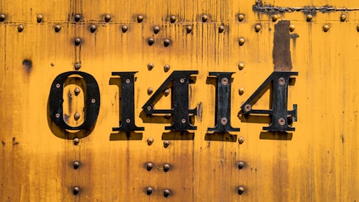 A vibrant yellow metal surface with multiple rivets, featuring the black painted numbers 01414. The background shows signs of wear and rust.