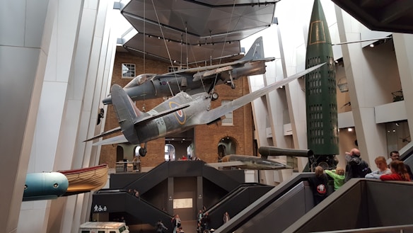 Interior of a museum featuring suspended aircraft and rockets with visitors observing from various levels and walkways. The setting includes modern architectural elements and a brick wall backdrop.