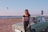 A young woman with long red hair stands in front of a vintage green car parked on a sandy area near a body of water. The sky is clear and kitesurfers can be seen in the distance over the water. The woman is wearing a black t-shirt with the word 'FEARLESS' printed on it in bold letters. Her hair is covering part of her face, suggesting a windy environment. A Union Jack flag is attached to the car's roof.