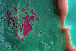 A textured surface with peeling green paint revealing a rusty red layer beneath. The paint is chipped and corroded in several areas, showcasing an irregular pattern of degradation. The surface has patches where the red and green mix, creating an abstract, artistic appearance.
