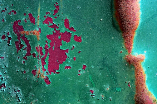 A textured surface with peeling green paint revealing a rusty red layer beneath. The paint is chipped and corroded in several areas, showcasing an irregular pattern of degradation. The surface has patches where the red and green mix, creating an abstract, artistic appearance.