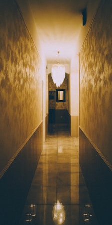 A dimly lit hallway with reflective tiled floors and softly textured walls. The focal point is a chandelier hanging from the ceiling, casting a warm, inviting glow along the corridor. At the end of the hallway, there is a mirror and some dark-toned furnishings adding depth to the space.