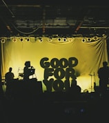 Silhouettes of musicians perform on stage with backlit letters spelling 'Good For You'. The lighting creates a dramatic contrast, highlighting the outlines of music equipment and band members.