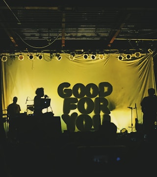 Silhouettes of musicians perform on stage with backlit letters spelling 'Good For You'. The lighting creates a dramatic contrast, highlighting the outlines of music equipment and band members.