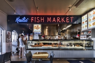 A vibrant fish market with a prominent neon sign reading 'Kailis' Fish Market'. There are people browsing the seafood selection displayed in a large glass counter filled with fresh seafood items. The interior features menu boards and food items decorated with holiday themes. The flooring is tiled and there are tables and chairs for seating.