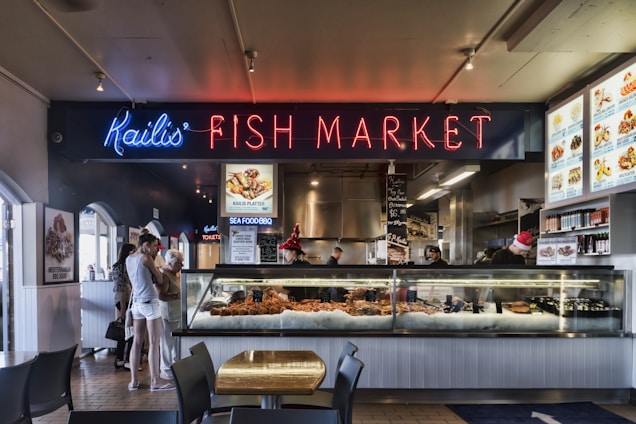A vibrant fish market with a prominent neon sign reading 'Kailis' Fish Market'. There are people browsing the seafood selection displayed in a large glass counter filled with fresh seafood items. The interior features menu boards and food items decorated with holiday themes. The flooring is tiled and there are tables and chairs for seating.