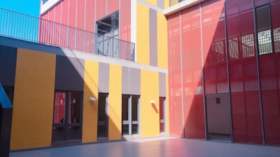 A modern architectural design featuring vibrant red and yellow sections on the exterior of a building. The area is illuminated by natural light, casting shadows on the textured walls. There are large glass windows and railings on the upper level.