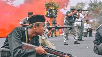 A group of people wearing military-style uniforms engage in a mock battle or reenactment. They are crouched on the street, holding rifles aimed forward. A dramatic red smoke billows in the background, adding intensity to the scene. Some participants wear distinctive caps or helmets, and a few have visible vintage equipment like backpacks. A flower arrangement can be seen behind them.