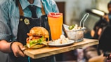 A person dressed in a striped shirt and apron is holding a wooden tray with a meal consisting of a hamburger with lettuce, tomato, and cheese. Beside the burger is a basket of fried snacks and a tall glass of orange juice with slices of fruit on top.