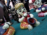 Colorful statuettes of musicians sit on a green surface. The figures are intricately painted with bold colors and have exaggerated facial features, wearing traditional attire with ornate patterns. Some figures are playing musical instruments, such as a drum and possibly a trumpet.