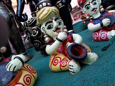 Colorful statuettes of musicians sit on a green surface. The figures are intricately painted with bold colors and have exaggerated facial features, wearing traditional attire with ornate patterns. Some figures are playing musical instruments, such as a drum and possibly a trumpet.