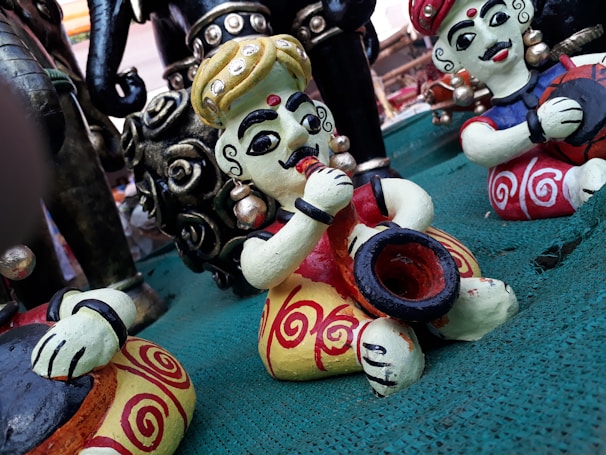 Colorful statuettes of musicians sit on a green surface. The figures are intricately painted with bold colors and have exaggerated facial features, wearing traditional attire with ornate patterns. Some figures are playing musical instruments, such as a drum and possibly a trumpet.
