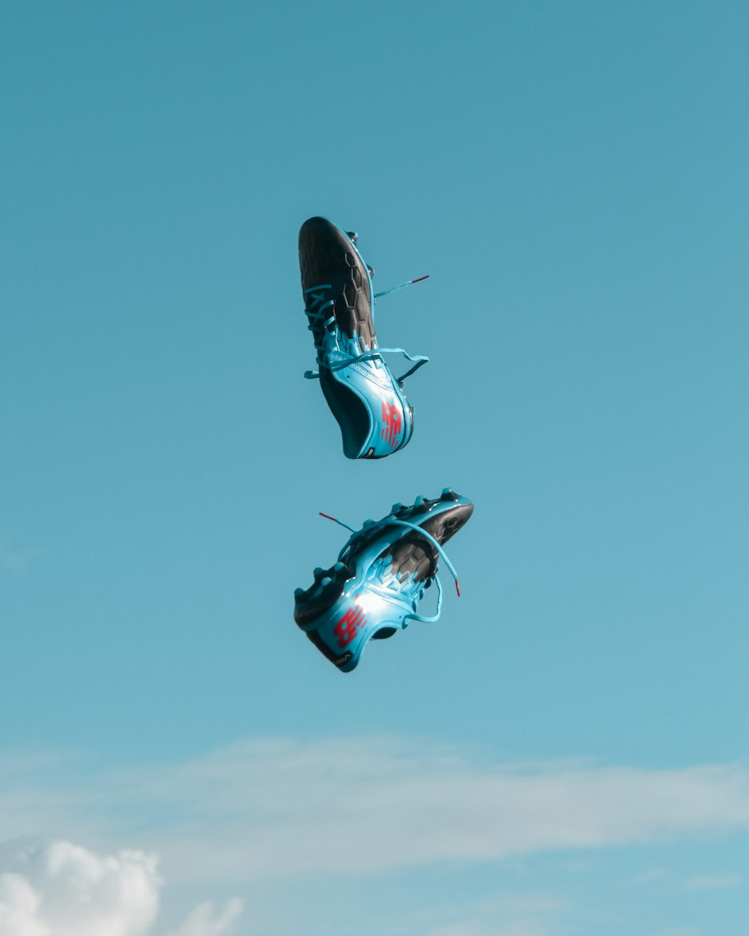 pair of blue-and-black New Balance cleats in the air under teal sky with white clouds