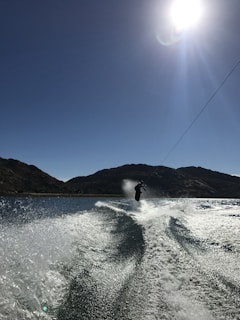 A person is wakeboarding on a clear body of water under bright sunlight. The water is creating a spray and visible wake trails as the boarder performs a jump or trick. The background features silhouetted hills under a clear blue sky with the sun shining brightly.