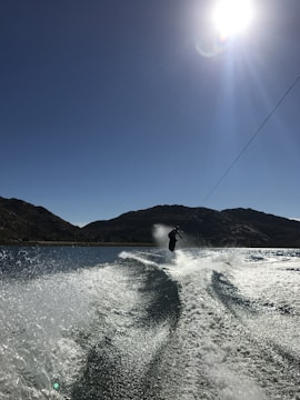 A person is wakeboarding on a clear body of water under bright sunlight. The water is creating a spray and visible wake trails as the boarder performs a jump or trick. The background features silhouetted hills under a clear blue sky with the sun shining brightly.