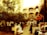 A bustling outdoor marketplace with multiple stalls lined with goods, surrounded by trees and an architectural backdrop featuring ornate archways and balconies. People are seen interacting, carrying goods, and browsing through items under the warm sunlight. The atmosphere is lively and busy, reflecting a traditional market scene.