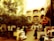A bustling outdoor marketplace with multiple stalls lined with goods, surrounded by trees and an architectural backdrop featuring ornate archways and balconies. People are seen interacting, carrying goods, and browsing through items under the warm sunlight. The atmosphere is lively and busy, reflecting a traditional market scene.