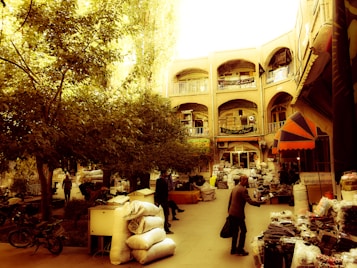 A bustling outdoor marketplace with multiple stalls lined with goods, surrounded by trees and an architectural backdrop featuring ornate archways and balconies. People are seen interacting, carrying goods, and browsing through items under the warm sunlight. The atmosphere is lively and busy, reflecting a traditional market scene.