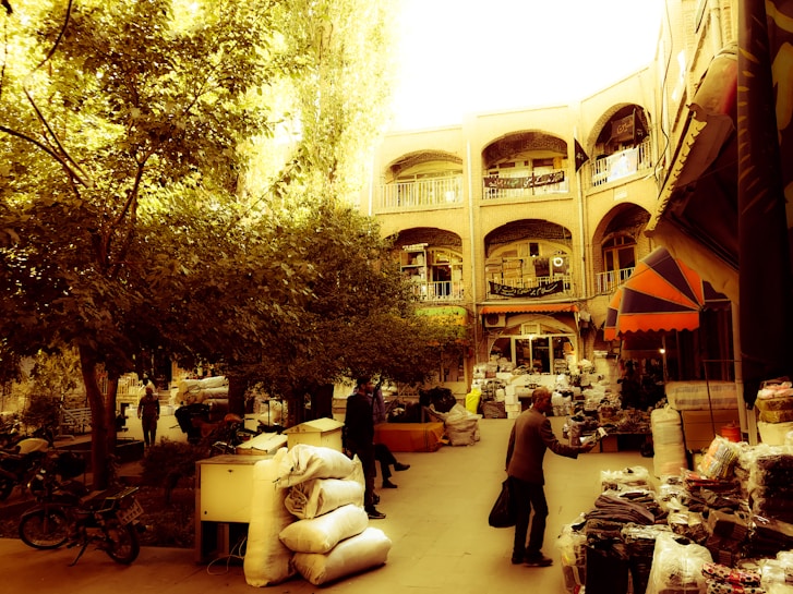 A bustling outdoor marketplace with multiple stalls lined with goods, surrounded by trees and an architectural backdrop featuring ornate archways and balconies. People are seen interacting, carrying goods, and browsing through items under the warm sunlight. The atmosphere is lively and busy, reflecting a traditional market scene.