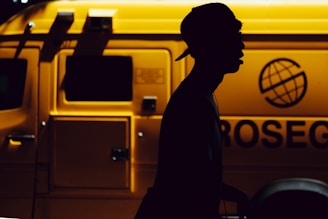 A silhouette of a person wearing a cap is prominently featured against the backdrop of a brightly lit yellow service vehicle with the word 'ROSEG' partially visible and a globe logo. The contrast creates a dramatic interplay between light and dark.
