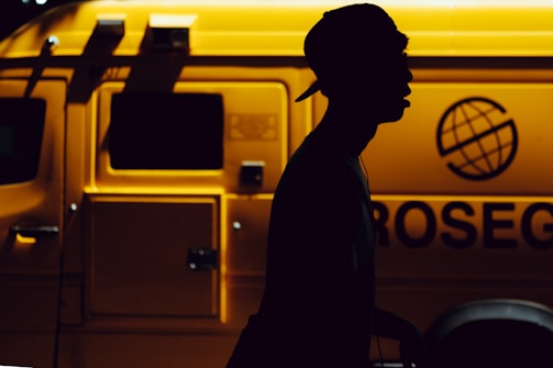A silhouette of a person wearing a cap is prominently featured against the backdrop of a brightly lit yellow service vehicle with the word 'ROSEG' partially visible and a globe logo. The contrast creates a dramatic interplay between light and dark.