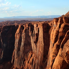 A vast, rugged canyon with steep, towering walls carved by natural forces. The layered rock formations display varying shades of brown, orange, and red, creating a stunning landscape. The horizon reveals a distant, hazy mountain range under a clear blue sky.