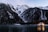 Snow-capped mountains rise steeply in the background, surrounded by dense, dark forests. At the base of the mountains, a tranquil lake reflects their majestic presence. A cozy, warmly lit cabin sits by the water's edge, its lights illuminating the surrounding trees.