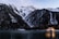 Snow-capped mountains rise steeply in the background, surrounded by dense, dark forests. At the base of the mountains, a tranquil lake reflects their majestic presence. A cozy, warmly lit cabin sits by the water's edge, its lights illuminating the surrounding trees.
