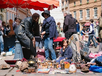 People are gathered at an outdoor market with various items displayed on the ground, including masks, books, and household objects. A child, surrounded by colorful textiles, sits near the items while multiple adults browse and interact. The scene is lively, with bright sunlight and the market setting in a bustling city square.