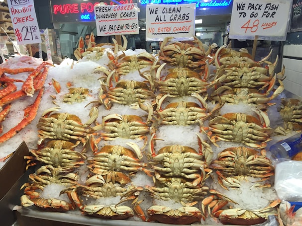 A market display featuring numerous Dungeness crabs arranged on a bed of ice. Signs advertising fresh cooked crabs, the price for halibut cheeks, and packaging services for take-out are visible. The setting appears vibrant and busy, typical of a seafood market.