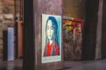 A stylized portrait of a person with long hair is displayed on a wooden panel in an urban setting. The artwork features a color scheme of red, blue, and beige with a bold message at the bottom reading 'We The People - Protect Each Other'. The background includes graffiti on metal structures and industrial elements, giving a gritty, artistic atmosphere.
