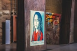 A stylized portrait of a person with long hair is displayed on a wooden panel in an urban setting. The artwork features a color scheme of red, blue, and beige with a bold message at the bottom reading 'We The People - Protect Each Other'. The background includes graffiti on metal structures and industrial elements, giving a gritty, artistic atmosphere.