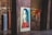 A stylized portrait of a person with long hair is displayed on a wooden panel in an urban setting. The artwork features a color scheme of red, blue, and beige with a bold message at the bottom reading 'We The People - Protect Each Other'. The background includes graffiti on metal structures and industrial elements, giving a gritty, artistic atmosphere.