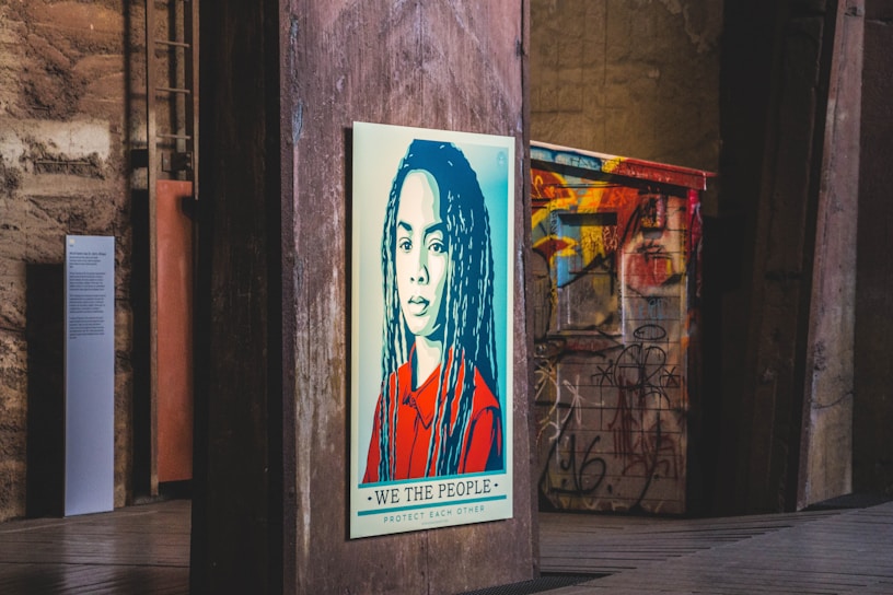 A stylized portrait of a person with long hair is displayed on a wooden panel in an urban setting. The artwork features a color scheme of red, blue, and beige with a bold message at the bottom reading 'We The People - Protect Each Other'. The background includes graffiti on metal structures and industrial elements, giving a gritty, artistic atmosphere.
